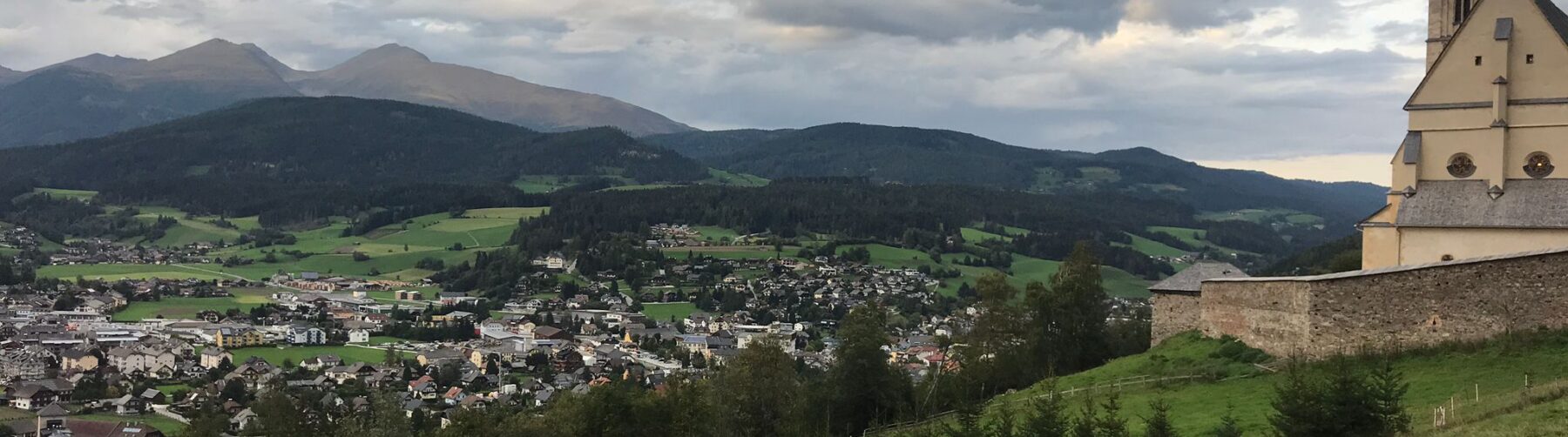 Von einem Hügel aus ist die Stadt Tamsweg im Lungau fotografiert. Im Vordergrund ist eine Wiese, rechts eine Kirche und im Hintergrund sind Berge. Der Himmel ist wolkig.