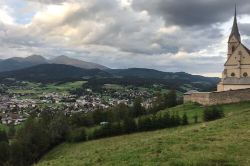 The city of Tamsweg in the region Lungau is shown from a hill. Behind Tamsweg, there are mountains and the sky is cloudy.